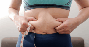 Person pinching abdominal belly fat while holding a measuring tape to track weight-loss progress. manage plateaus in your weight loss journey